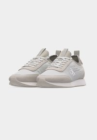 Ζευγάρι γκρι και άσπρα αθλητικά παπούτσια χαμηλού ύψους με κορδόνια και λογότυπο "cK" στην εξωτερική πλευρά, ελαφρώς γυρισμένα προς τα δεξιά σε απλό φόντο.