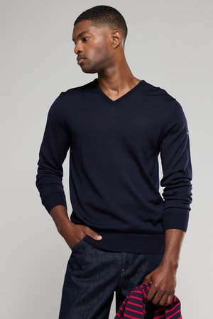 Marineblauer V-Ausschnitt-Pullover aus weichem Material mit gerippten Bündchen und Saum, kombiniert mit dunklen Jeans. Ein gestreiftes Hemd liegt über einem Arm.