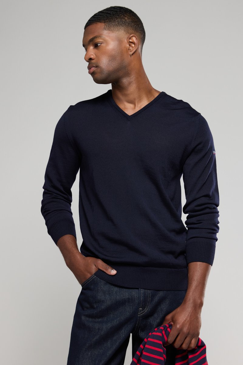 Marineblauer V-Ausschnitt-Pullover aus weichem Material mit gerippten Bündchen und Saum, kombiniert mit dunklen Jeans. Ein gestreiftes Hemd liegt über einem Arm.