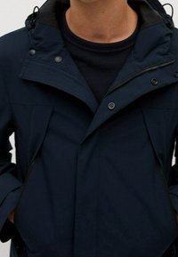 Tmavě modrá bunda se zipem a zapínáním na druky, s nastavitelnou kapucí a bočními kapsami. Vyrobena z hladké tkaniny se strukturovaným designem.