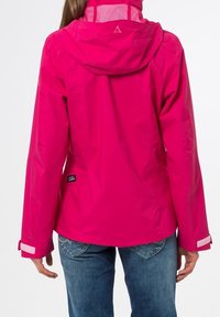 Donna con una giacca a tre quarti fucsia e jeans blu, vista da dietro su uno sfondo bianco uniforme.