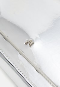 Sömlös, silverfärgad metallic clutchväska med slät yta, som har en subtil präglad logotyp och en dekorativ nitar trim längs kanten.