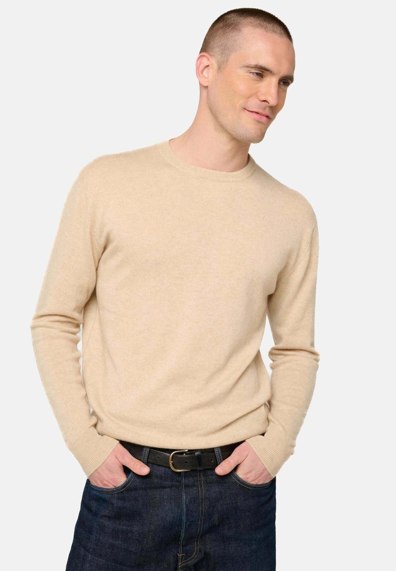 Pull en maille légère couleur beige, avec des manches longues et des poignets côtelés, associé à un jean en denim foncé et une ceinture noire.