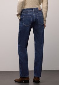 Pantalones vaqueros de mezclilla azul oscuro con corte recto, que cuentan con bolsillos traseros y un parche de cuero en la cintura. Combinados con un suéter beige y botas marrones.