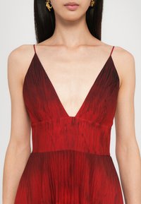 Punane ilma varrukateta kleit, millel on sügav V-kaelus ja tekstuuriga, volditud disain. Kanga pinnal on gradient muster, kus tumedamad aktsendid asuvad üleval.