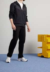 Black zip-up hoodie with patterned upper panel, worn over a white t-shirt, paired with black pants and white athletic shoes. Yellow storage unit nearby.