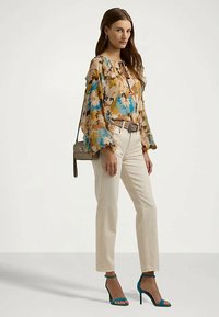 Donna che indossa una camicetta floreale a maniche lunghe, pantaloni beige a vita alta, cintura con motivo, sandali alti teal e porta una piccola borsa a tracolla.