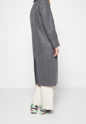 Personne portant un long manteau gris, un pantalon blanc à jambes larges et des baskets colorées, debout de profil sur un fond blanc uni.