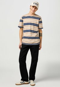 Camiseta a rayas beige con líneas en azul marino y blanco, pantalones negros y zapatillas blancas con rayas en azul marino. El sombrero dice "Una leyenda en vaqueros."