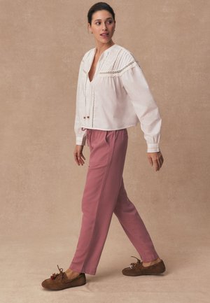 Donna che cammina indossando una camicetta bianca a maniche lunghe, pantaloni rosa e scarpe in stile mocassino marrone su uno sfondo beige.