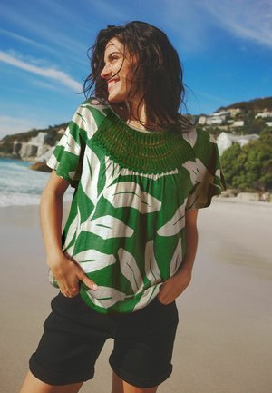 Grünes Kurzarmshirt mit großen weißen Blattmustern und einem strukturierten, grünen, gestickten Kragen, kombiniert mit schwarzen Shorts. Strandkulisse.