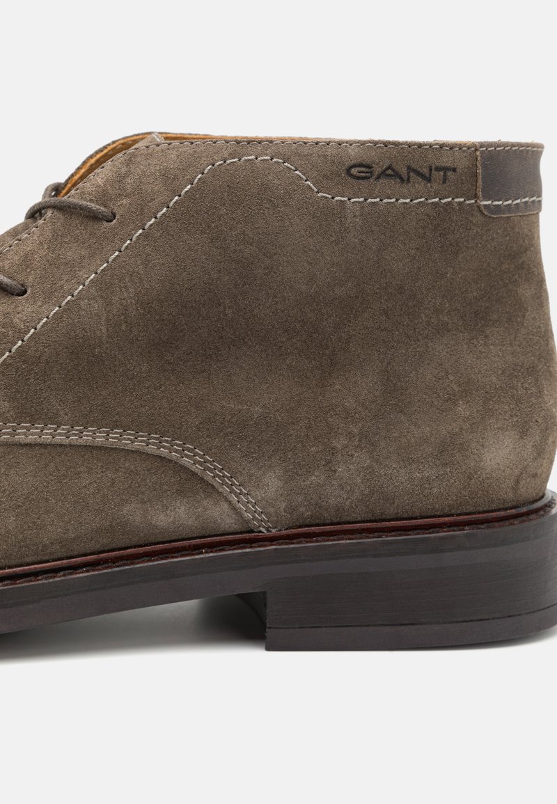 Brązowy zamszowy botek za kostkę z przodem na sznurki, kontrastowymi szwami, ciemną podeszwą i wytłoczonym logo GANT w pobliżu pięty.
