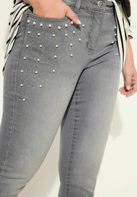 Jeans de mezclilla gris con adornos de perlas en el bolsillo delantero, con un corte ajustado y un ligero desgastado para dar textura.