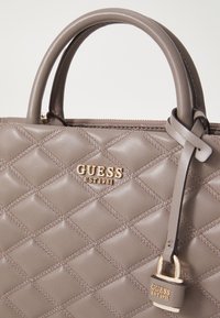 Bolso acolchado gris con acabado brillante, logo dorado "GUESS EST 1981", dobles asas y una correa pequeña con detalle de hardware.