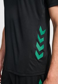 Czarna sportowa koszulka o gładkiej fakturze, z krótkimi rękawami i zielonymi grafikami w kształcie szewronów po lewej stronie.