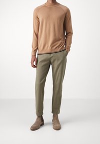 Beiger Strickpullover mit hohem Kragen, kombiniert mit olivgrünen, schmal geschnittenen Hosen. Hellbraune Wildleder-Stiefeletten runden das Outfit ab.