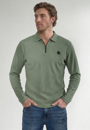 Homme aux cheveux bruns portant un polo vert à manches longues zippé et un jean bleu, debout les mains dans les poches, regardant à gauche.