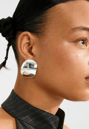 Donna con la pelle liscia che indossa un colletto nero a righe e un grande orecchino d'argento lucido e di forma irregolare all'orecchio sinistro, con i capelli raccolti.