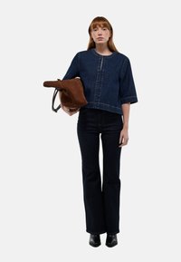 Veste en jean noir coupée avec des manches larges, associée à un jean flare noir. Le modèle tient un sac en suédine marron. Texture lisse et lignes épurées.