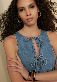 Top de denim con cierre de lazo, escote redondo y diseño sin mangas. Los accesorios incluyen pendientes de aro multicolores y pulseras apiladas.