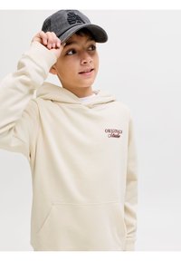 Αγόρι που φοράει ένα μπεζ φούτερ με κουκούλα με το κείμενο "Originals Studio", ρυθμίζοντας ένα σκουρόγρι baseball καπέλο, κοιτώντας ελαφρώς προς τα επάνω με φόντο απλό.