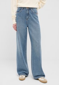 Persona con jeans a gamba larga di colore azzurro chiaro, sneakers beige e bianche, e una maglietta a maniche lunghe color crema, in piedi su uno sfondo semplice.