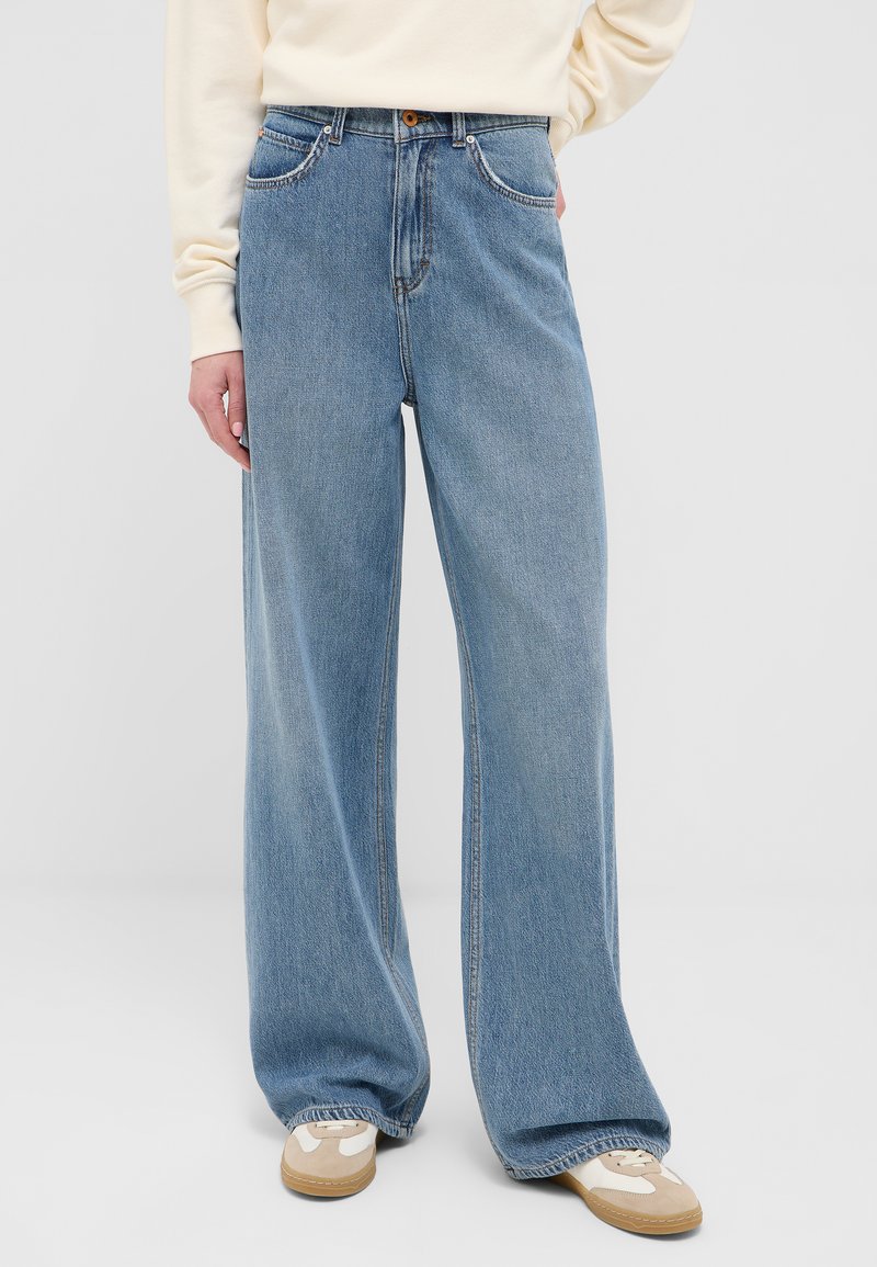 Persona con jeans a gamba larga di colore azzurro chiaro, sneakers beige e bianche, e una maglietta a maniche lunghe color crema, in piedi su uno sfondo semplice.