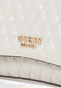 Bela prešita torbica iz sijočega materiala z zlatim logotipom "GUESS". Ima diamantski vzorec in fino šivanje ob robovih.