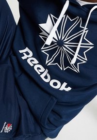 Felpa blu navy con logo Reebok bianco e grafica a stella. Presenta lacci bianchi e un piccolo accento del logo sull'orlo inferiore. Tessuto dalla consistenza morbida.