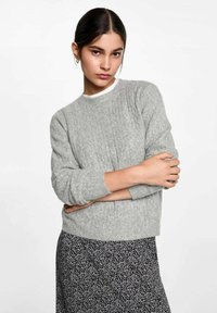 Světle šedý pletený svetr s ribbovanou strukturou, kulatým výstřihem a dlouhými rukávy, v kombinaci s černou sukní s květinovým vzorem.