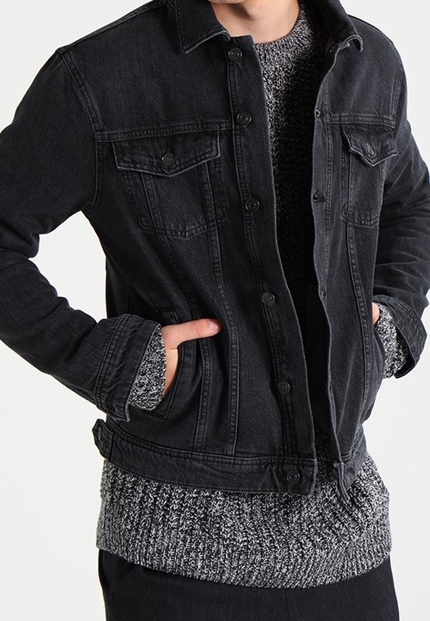 Personne portant une veste en denim noire par-dessus un pull en maille gris, les mains partiellement dans les poches de la veste.