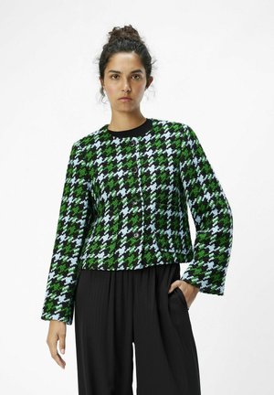 Chaqueta de espiga texturizada en verde, negro y azul claro, con cuello redondo, mangas largas y cinco botones en el frente. Combinada con pantalones negros.