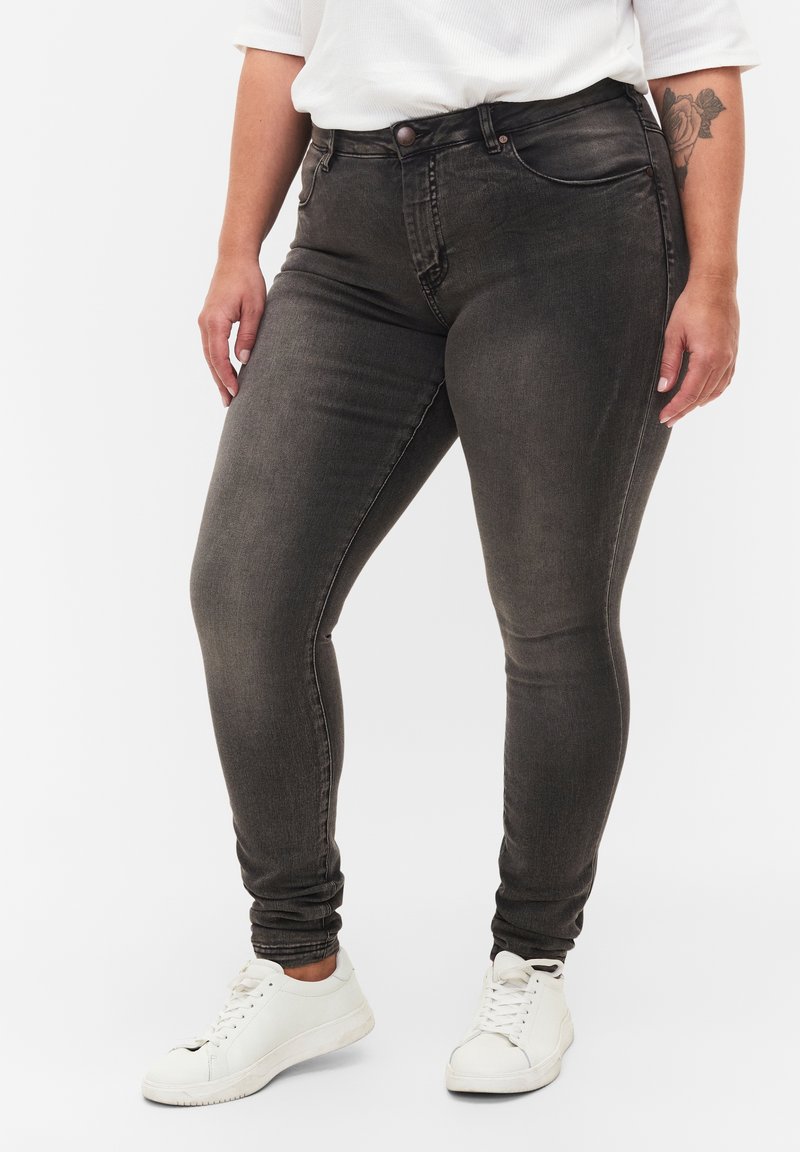 Personne portant un jean gris foncé ajusté et des baskets blanches, debout avec un haut blanc partiellement visible et un tatouage floral sur l'avant-bras gauche.