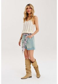 Biela pletená tielko s výstrihom do V, svetlé džínsy šortky a kvetinová šatka zviazaná okolo pása. Hnedé voľné čižmy po kolená.