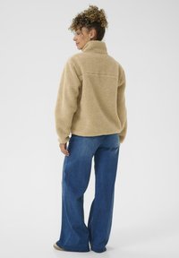 Pull en polaire beige avec un col montant, surface texturée et coupe ample, associé à un jean en denim bleu à jambes larges et des chaussures beiges.