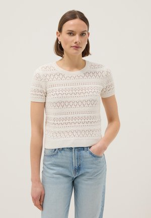 Donna che indossa un maglione bianco a maniche corte con motivi a cuore e diamante, jeans azzurri, in piedi con una mano in tasca.