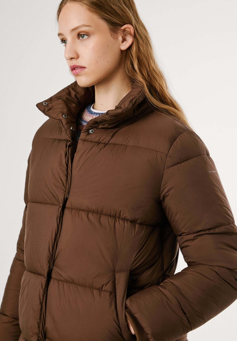 Młoda kobieta w brązowej kurtce puchowej z wysokim kołnierzem, stojąca z jedną ręką w kieszeni, patrząca w bok na jednolitym tle.