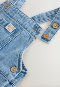 Vaaleansiniset farkkuhaalarit puisilla nappeilla ja pienellä kissan kasvokuvalla etutaskussa, aseteltuna tasaiselle valkoiselle pinnalle.