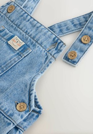 Világoskék farmer kertésznadrág fa gombokkal és egy apró macskafej folttal az első zsebén, fehér felületen kiterítve.