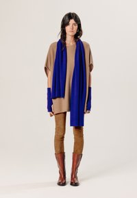 Bruine oversized gebreide top gecombineerd met bruine slim-fit broek, blauwe sjaal en bruine leren kniehoog laarzen. Inclusief vingerloze wanten in blauw.