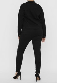 Vrouw in een fitted zwarte denim jack, zwarte skinny jeans en zwarte hoge hakken, vanachter gezien tegen een effen achtergrond.