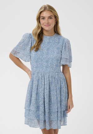 Młoda kobieta z długimi blond włosami nosi niebieską, kwiatową sukienkę z krótkim rękawem i warstwami, stojąc z jedną ręką na biodrze na jednolitym tle.