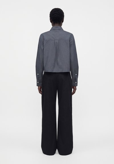 Personne vue de dos portant une chemise grise à fines rayures, courte, à manches longues, et un pantalon noir ample à jambes larges.