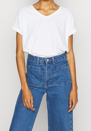 Personne portant un t-shirt blanc à manches courtes et col en V, rentré dans un jean bleu taille haute avec des poches plaquées à l'avant.