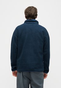 Νεαρό άτομο που φοράει ένα φλις μπουφάν σε σκούρο μπλε και γκρι παντελόνι, στέκεται με την πλάτη στραμμένη σε απλό λευκό φόντο.