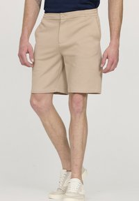 Shorts beige en tissu doux, avec une taille plate, une ceinture élastique et des poches latérales. Associés à des baskets de couleur claire.