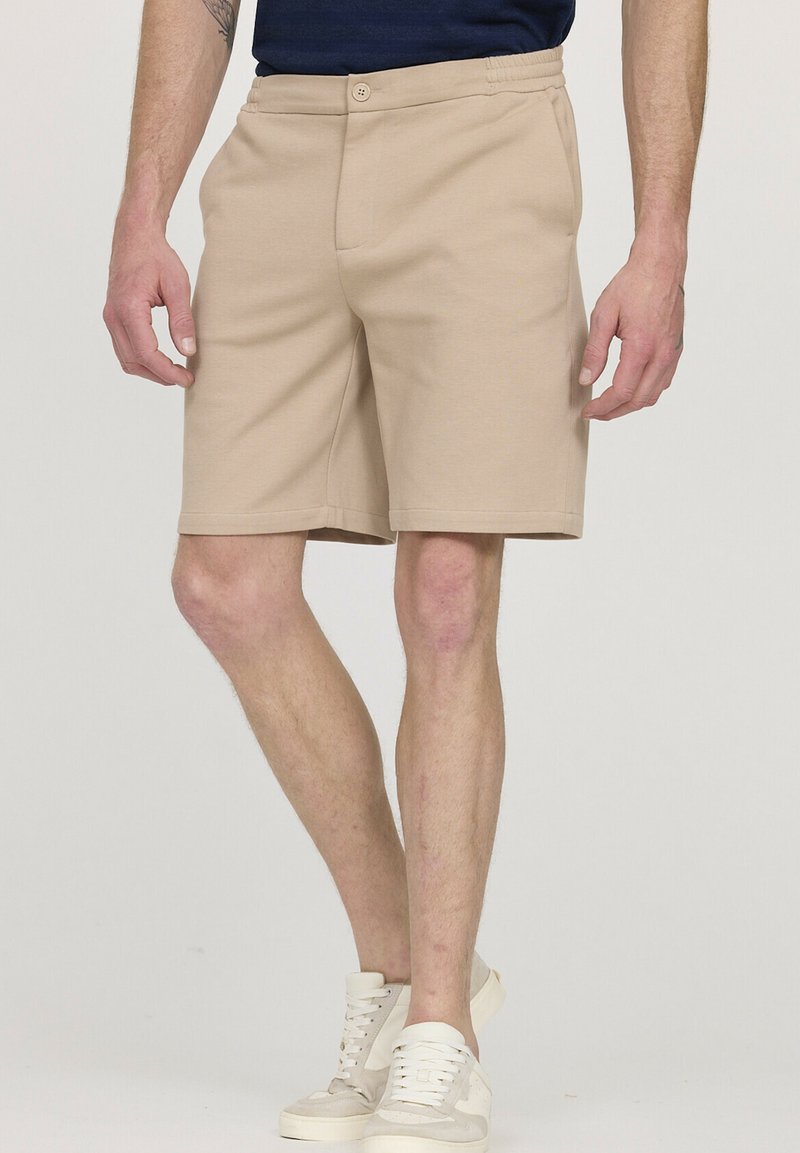 Shorts beige en tissu doux, avec une taille plate, une ceinture élastique et des poches latérales. Associés à des baskets de couleur claire.