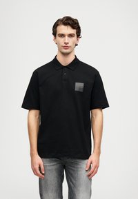 Svart polo t-shirt i bomull, med knäppt krage, korta ärmar och en grå fyrkantig logotyp på bröstet. Bärs med gråjeans.