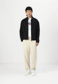 Svart teksturert jakke med glidelås foran over en hvit grafisk t-skjorte. Kombinert med lys beige bukser og hvite sneakers.