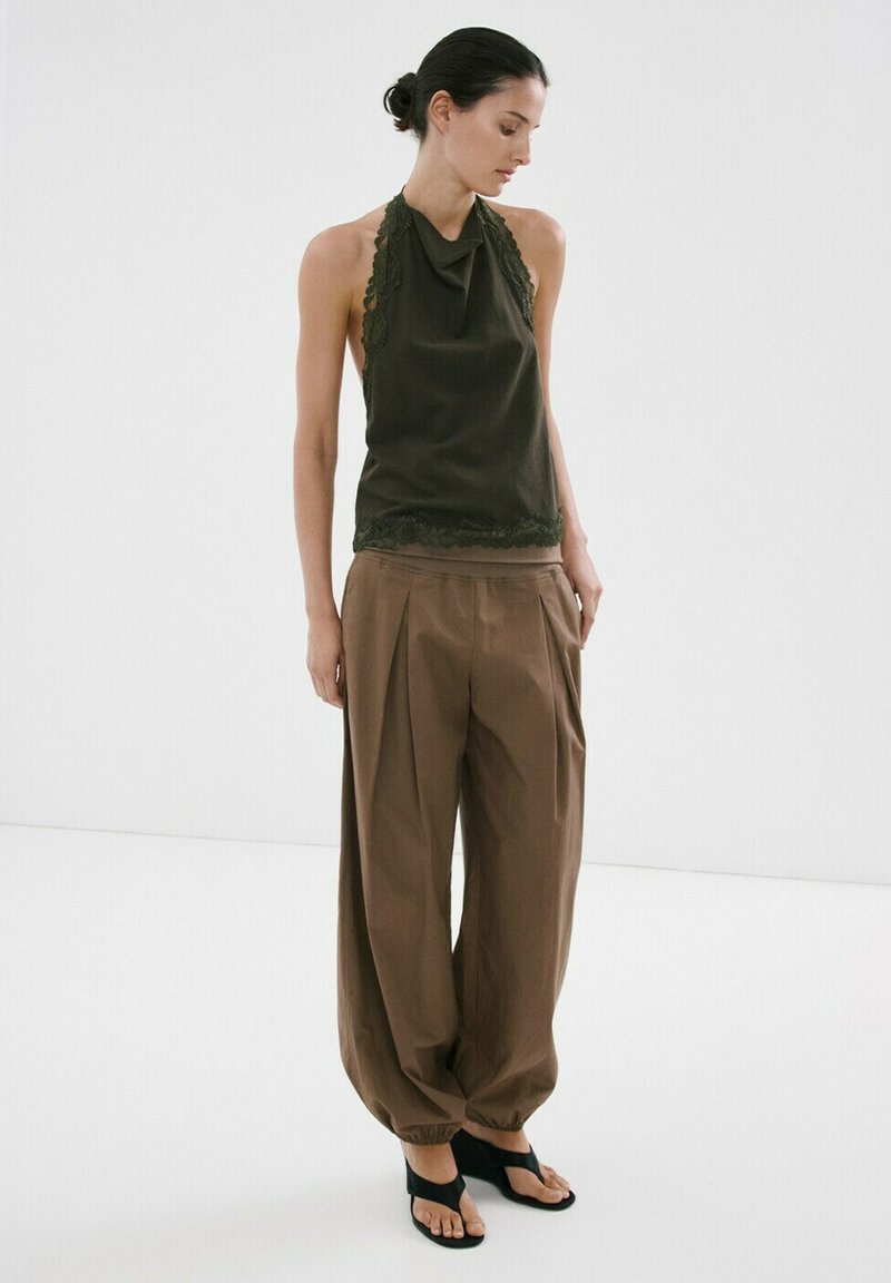 Femme portant un haut dos nu vert foncé orné de dentelle, un pantalon plissé marron ample et des sandales noires, debout devant un fond blanc.